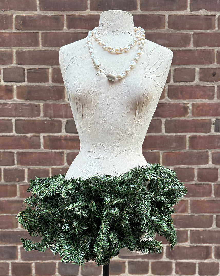 Mannequin wearing a green festive skirt and a pearl necklace with silver clasps against a brick wall