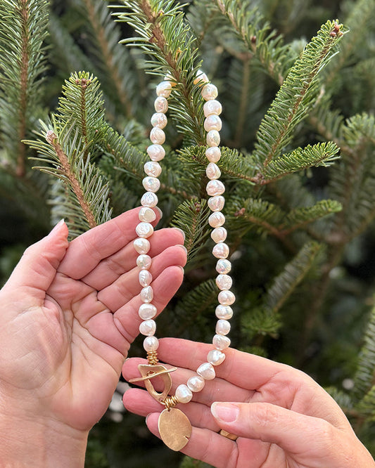Person holding a pearl necklace with a gold charm against a green tree background