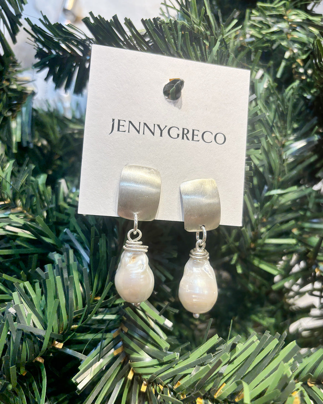 Earrings with pearls suspended from a silver metal square on a Jenny Greco tag against green foliage.
