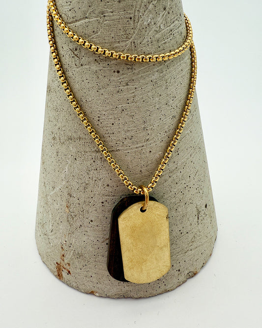 Gold necklace with a dog tag pendant on a textured gray stand.