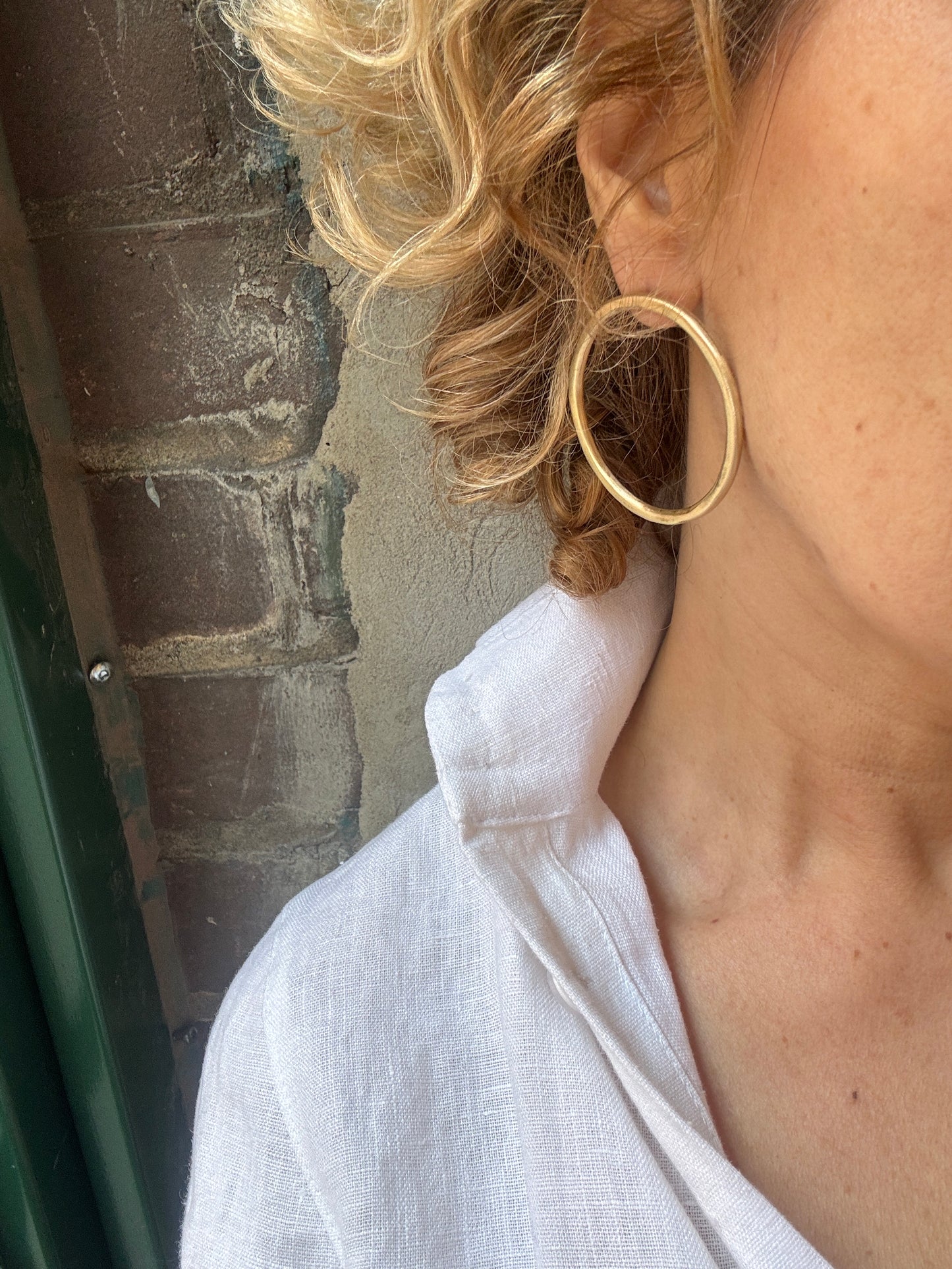 Close-up of a person with blonde hair and a white shirt wearing a gold hoop earrings standing against a brick wall.