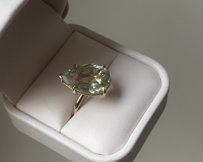 Gold ring with a green pearl shaped gemstone in a white ring box. 