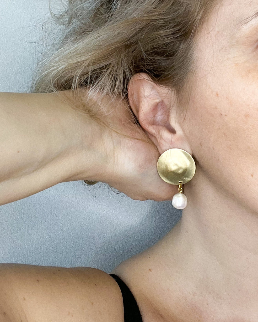 Close-up of a person wearing gold earrings with a pearl accent against a light blue background