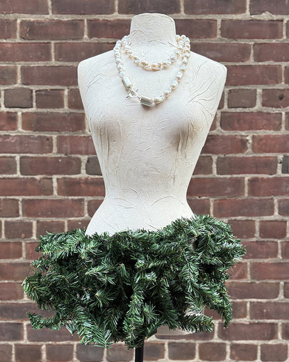 Mannequin wearing a green festive skirt  and a pearl necklace with silver clasps against a brick wall