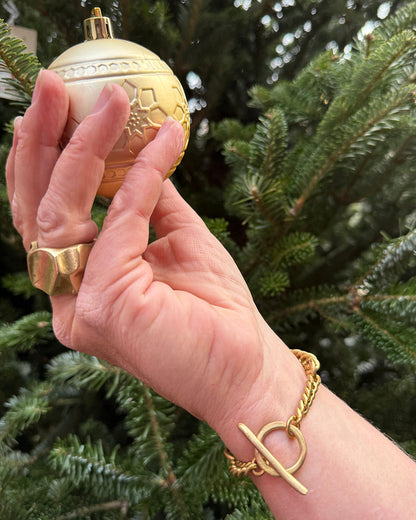 Hand holding a gold Christmas ornament with a tree in the background wearing a gold bracelet and ring. 