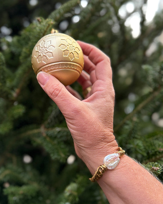 Hand holding a gold Christmas ornament against a blurred green tree background wearing a gold bracelet with a large pearl.