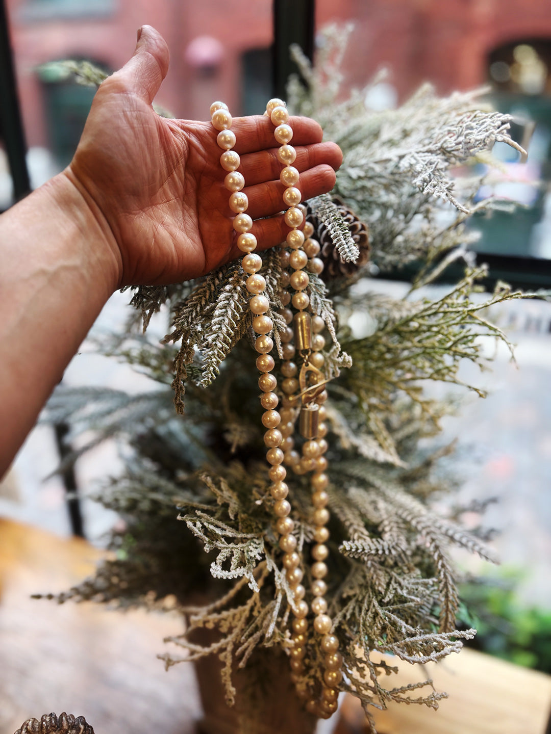 Hand holding a string of pearls over a decorative arrangement with greenery