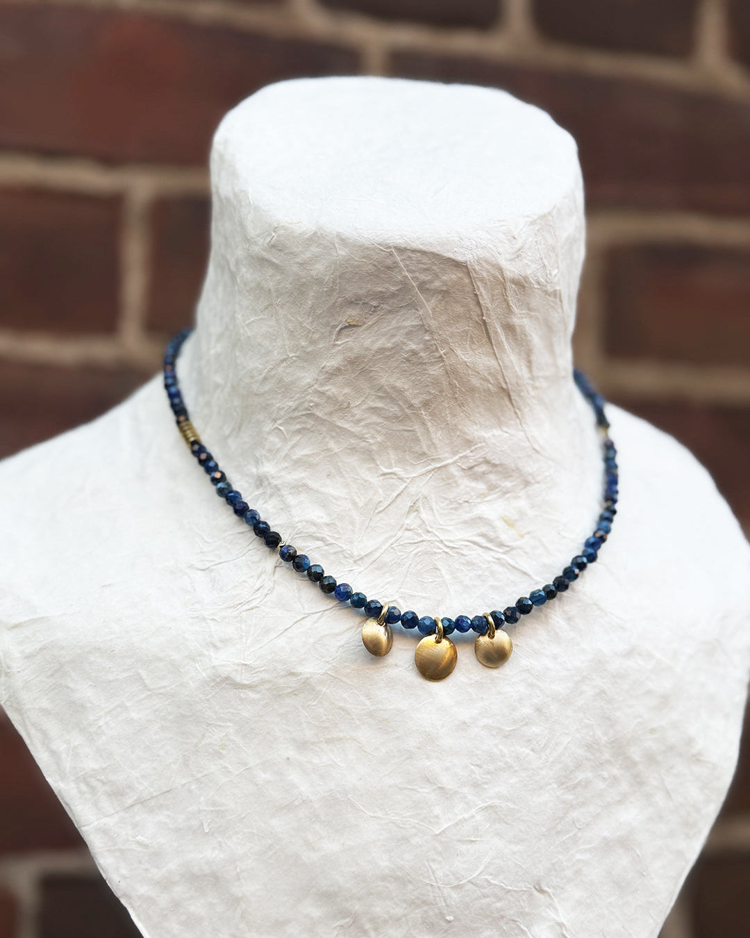 Necklace with blue beads and gold accents on a mannequin against a brick wall.