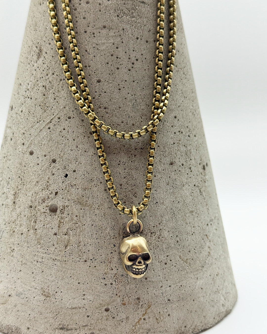 Gold chain necklace with a skull pendant on a textured gray stand.