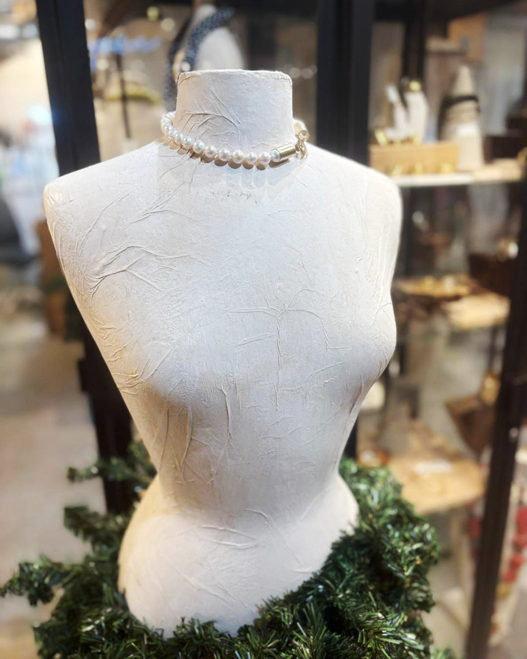 Mannequin head with a necklace on a mannequin, surrounded by greenery in an indoor setting.