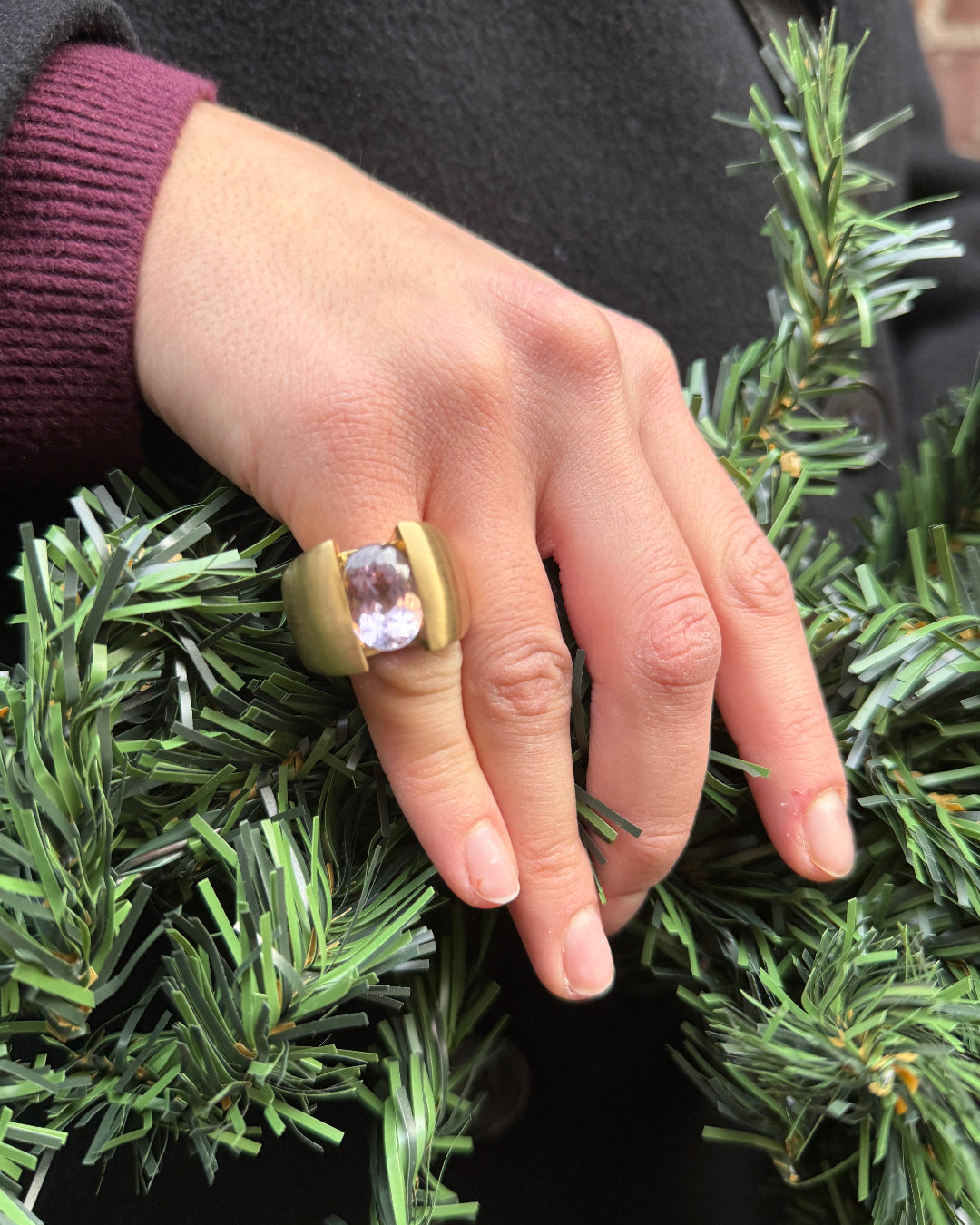Hand wearing a ring with a pink gemstone on green foliage