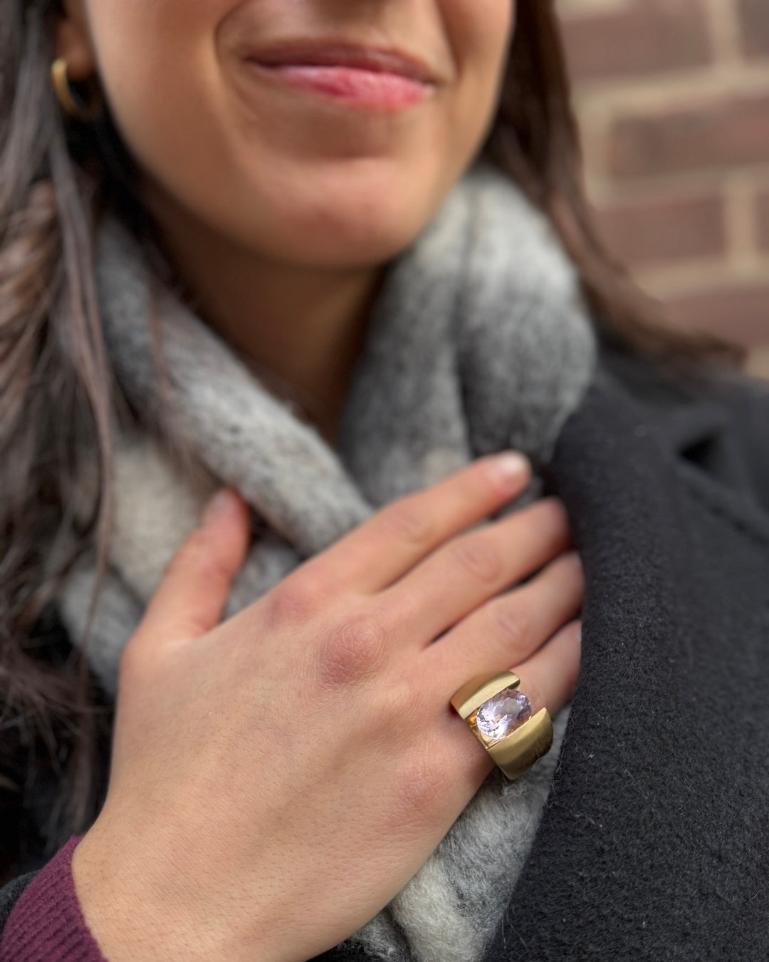 Woman wearing a gold ring with a purple gemstone, holding her hand to her chest against a brick wall.