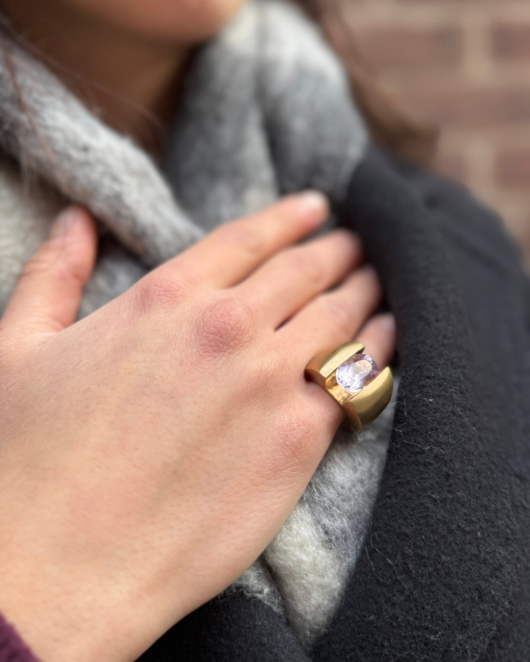 Hand wearing a gold ring with a diamond, holding a gray scarf against a brick wall.