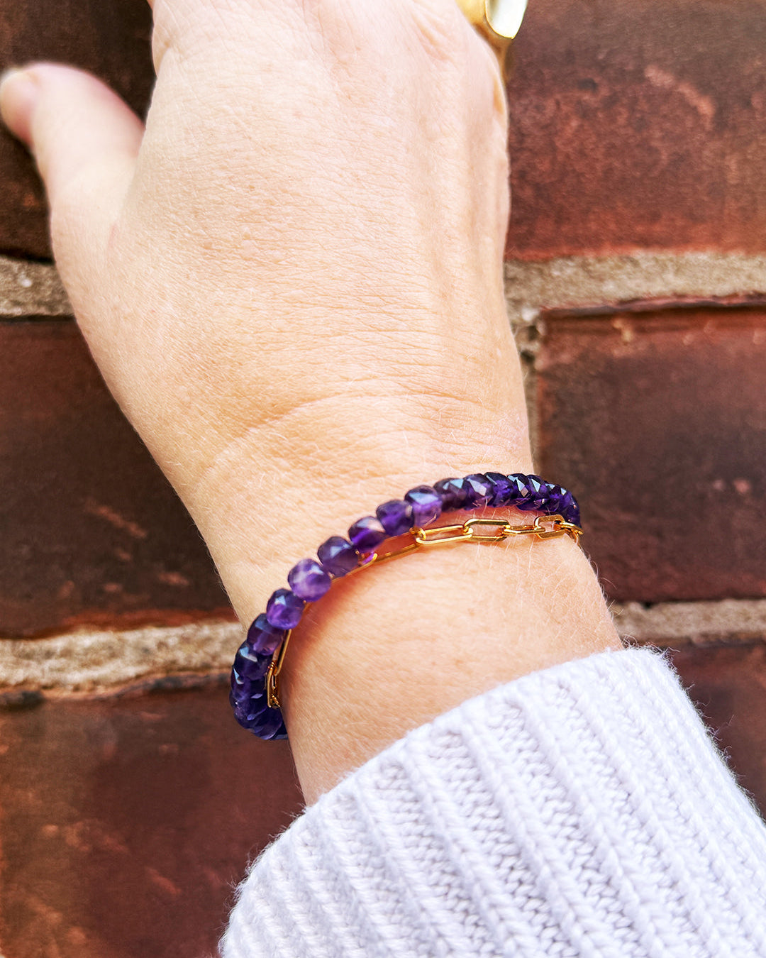 Hand wearing a purple beaded bracelet with gold accents against a brick wall background
