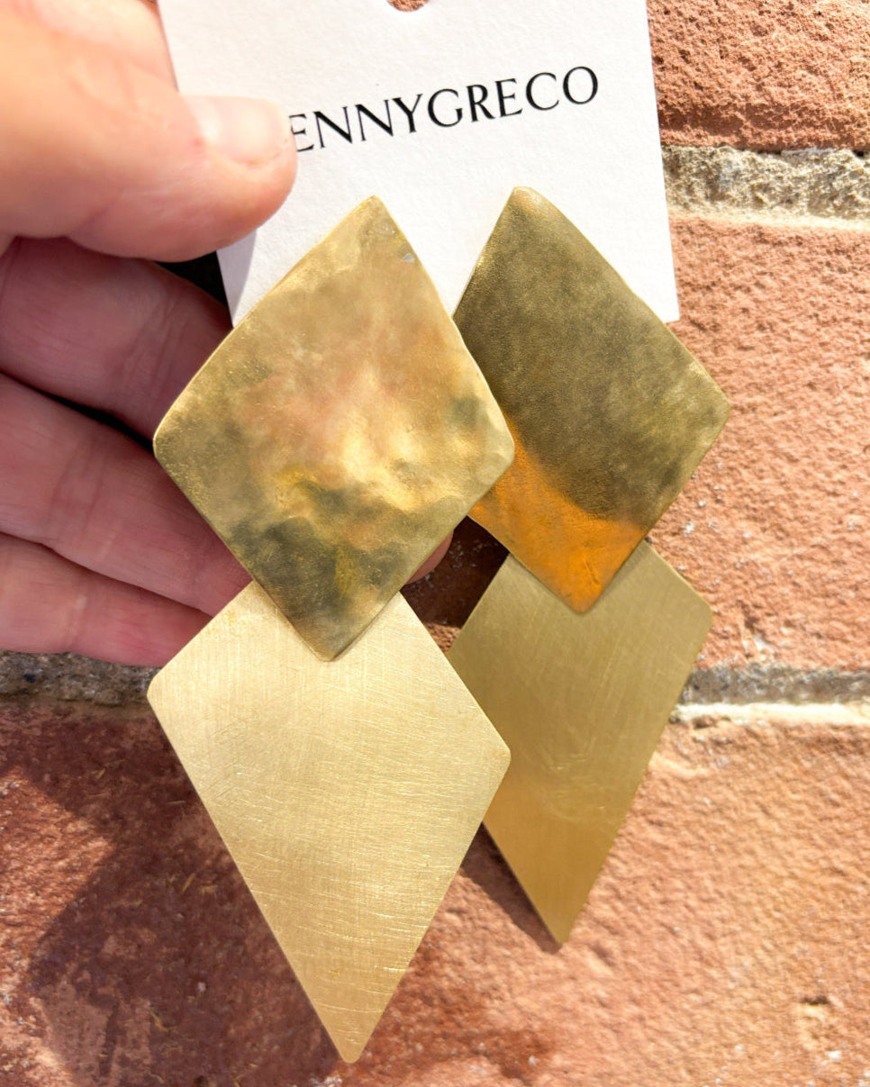 Gold geometric earrings held against a brick wall with 'JENNYGRECO' card.