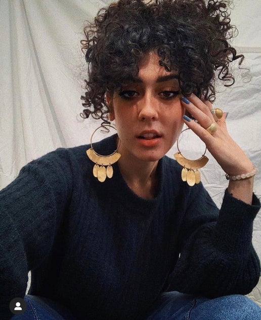 Woman wearing large gold coloured hoop earrings sitting against a neutral background