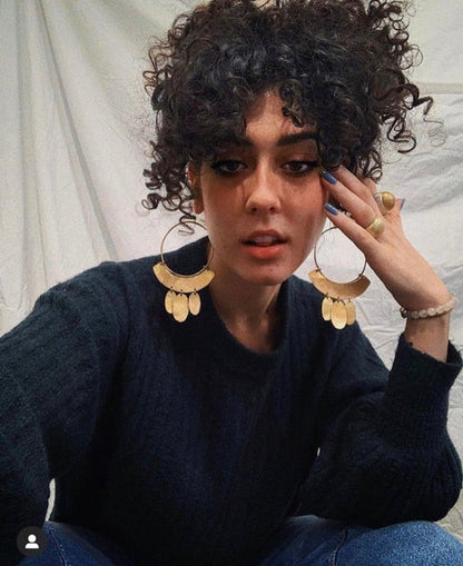 Woman wearing large gold coloured hoop earrings sitting against a neutral background