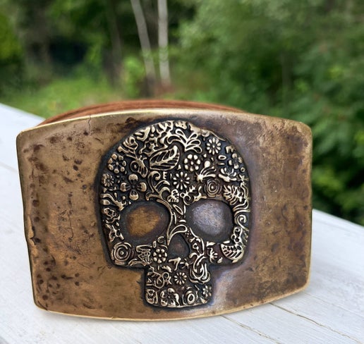 Bronze skull-shaped belt buckle on a white surface with a blurred green background