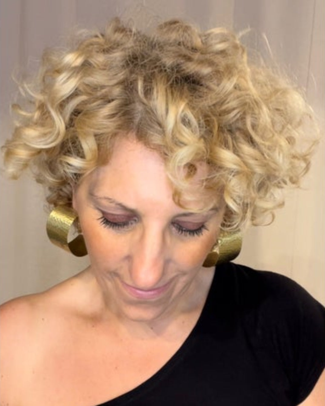Woman with curly blonde hair wearing chunky bronze hoop earrings and  wearing aa black top.