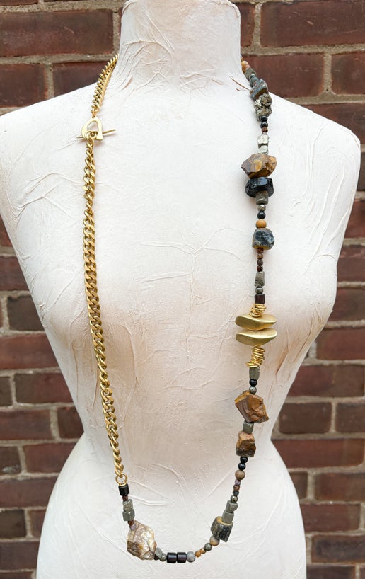 Mannequin wearing a necklace with gold chain and stone beads against a brick wall.