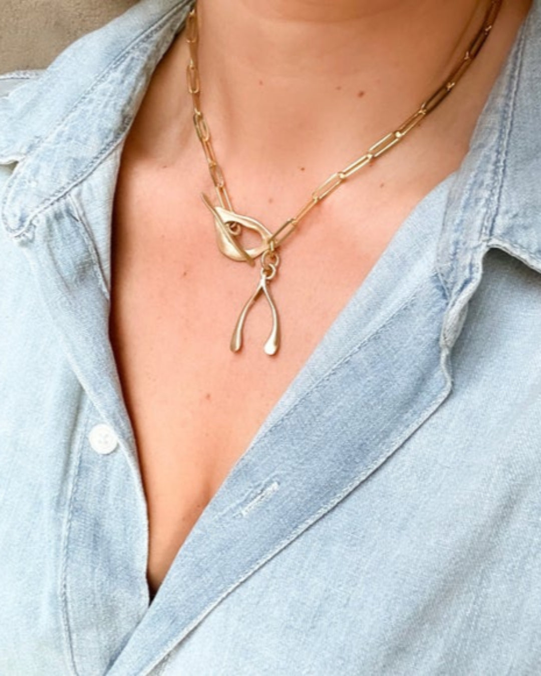 Gold necklace with a wishbone pendant worn by a person in a light blue denim shirt.