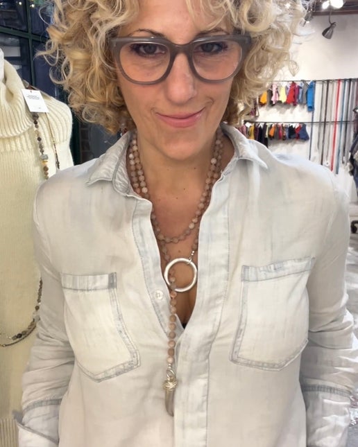 Woman wearing a light-colored shirt, glasses and a necklace with pink beads and silver accents in a clothing store setting