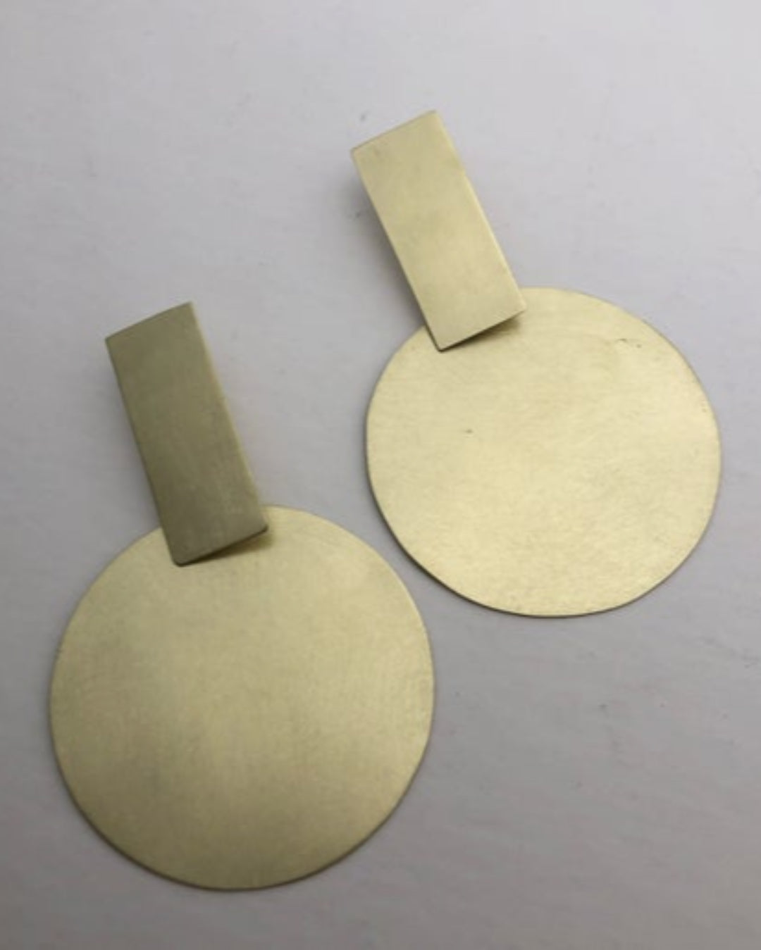 Two gold earrings with circular pendants and rectangular attachments on a gray background