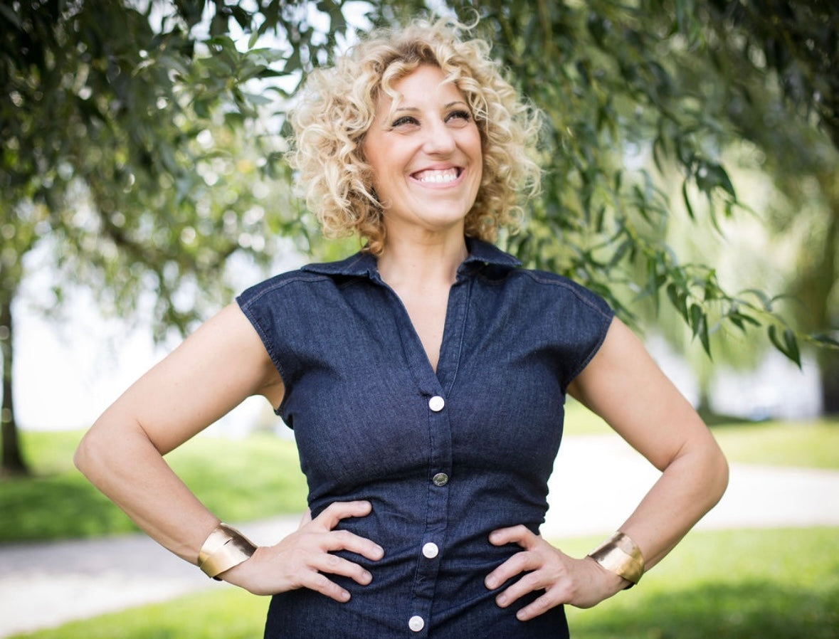 Woman in a denim dress  and two bronze cuffs standing outdoors with trees in the background