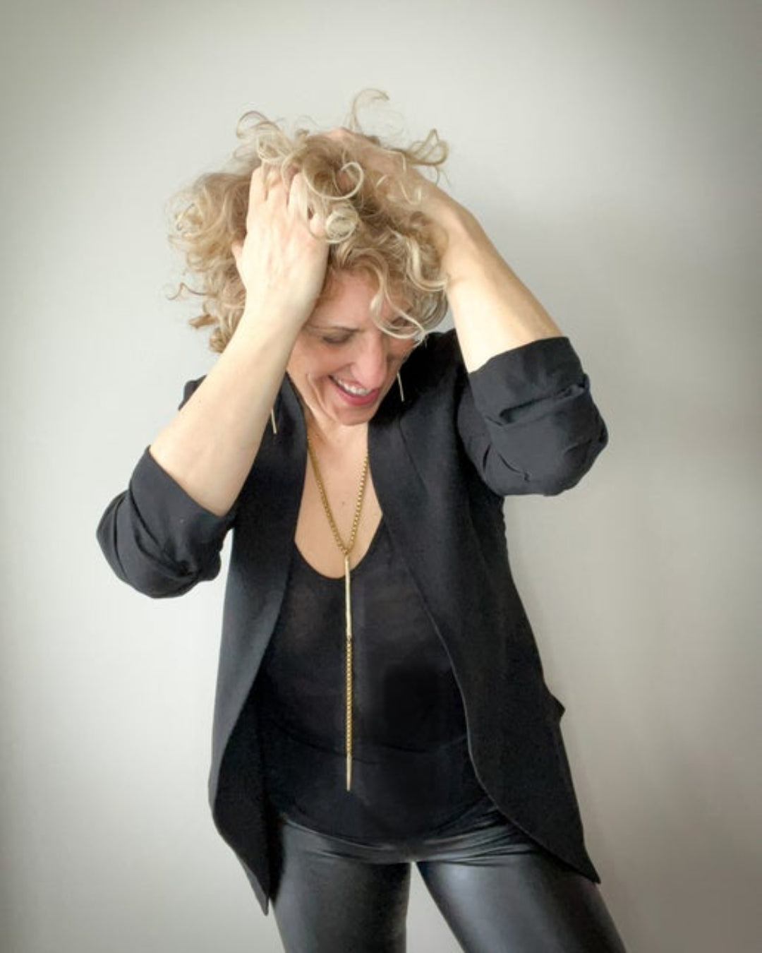 Woman with curly hair wearing a black outfit and gold necklace with bar details against a plain background