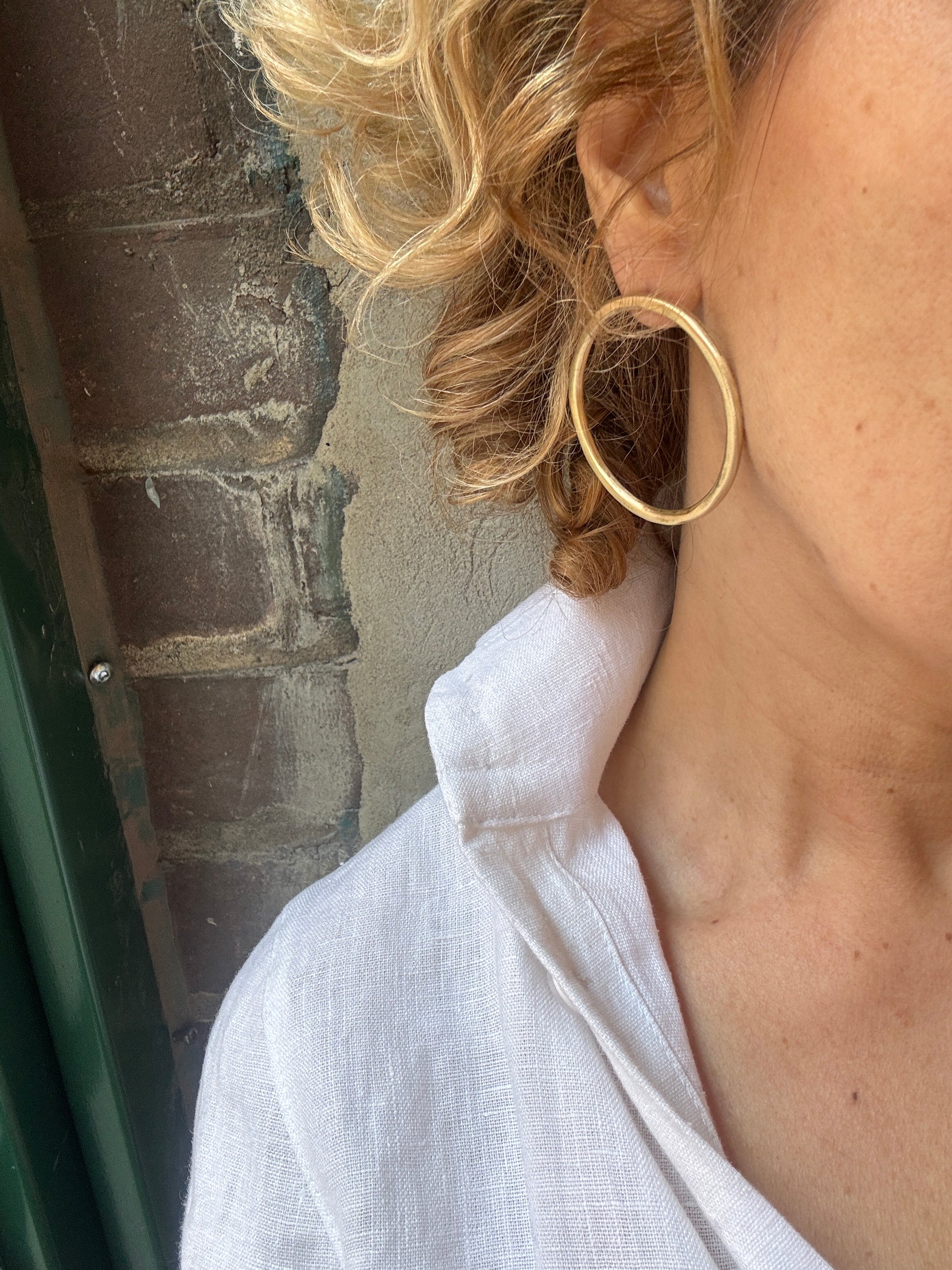 Close-up of a person with blonde hair and a white shirt wearing a gold hoop earrings standing against a brick wall. 