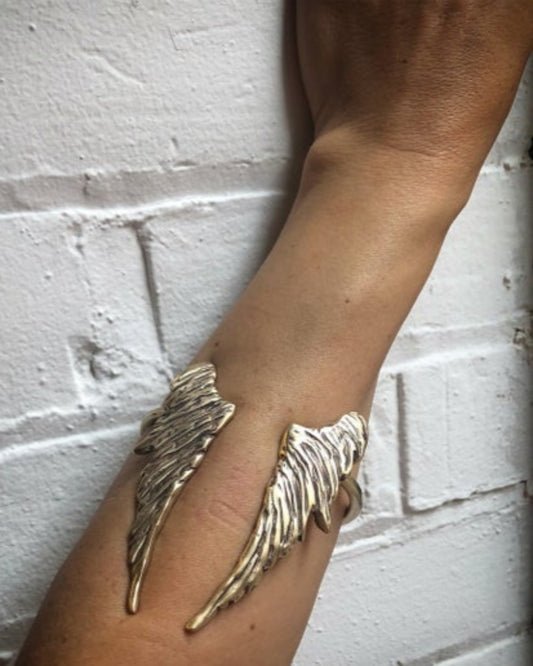 Gold cuff with wing detail on a wrist against a white brick wall.