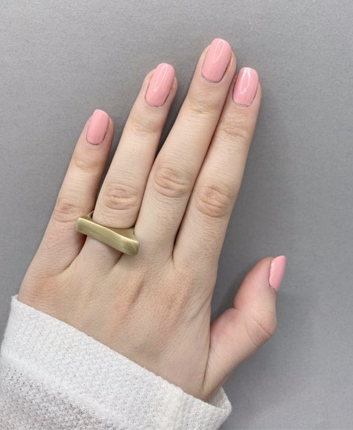 Hand wearing a geometric gold ring with pink nail polish on a grey background