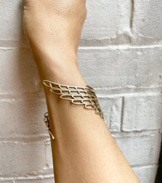 Gold bracelet with wing detail on a wrist against a light brick wall background