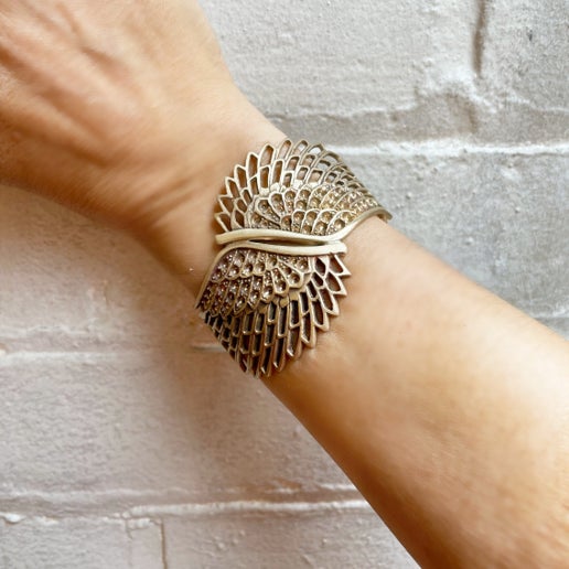 Bronze cuff with wing detailing on a wrist against a white brick background  