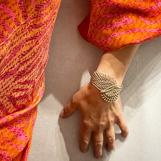 Hand wearing a gold cuff with wing detailing and an orange and pink patterned fabric on a white background 