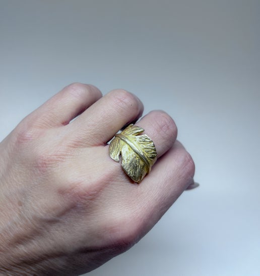 Gold feather-shaped ring on a person's finger against a plain background
