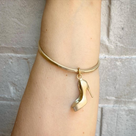 Gold bangle with a high-heeled shoe charm on a person's wrist against a neutral background