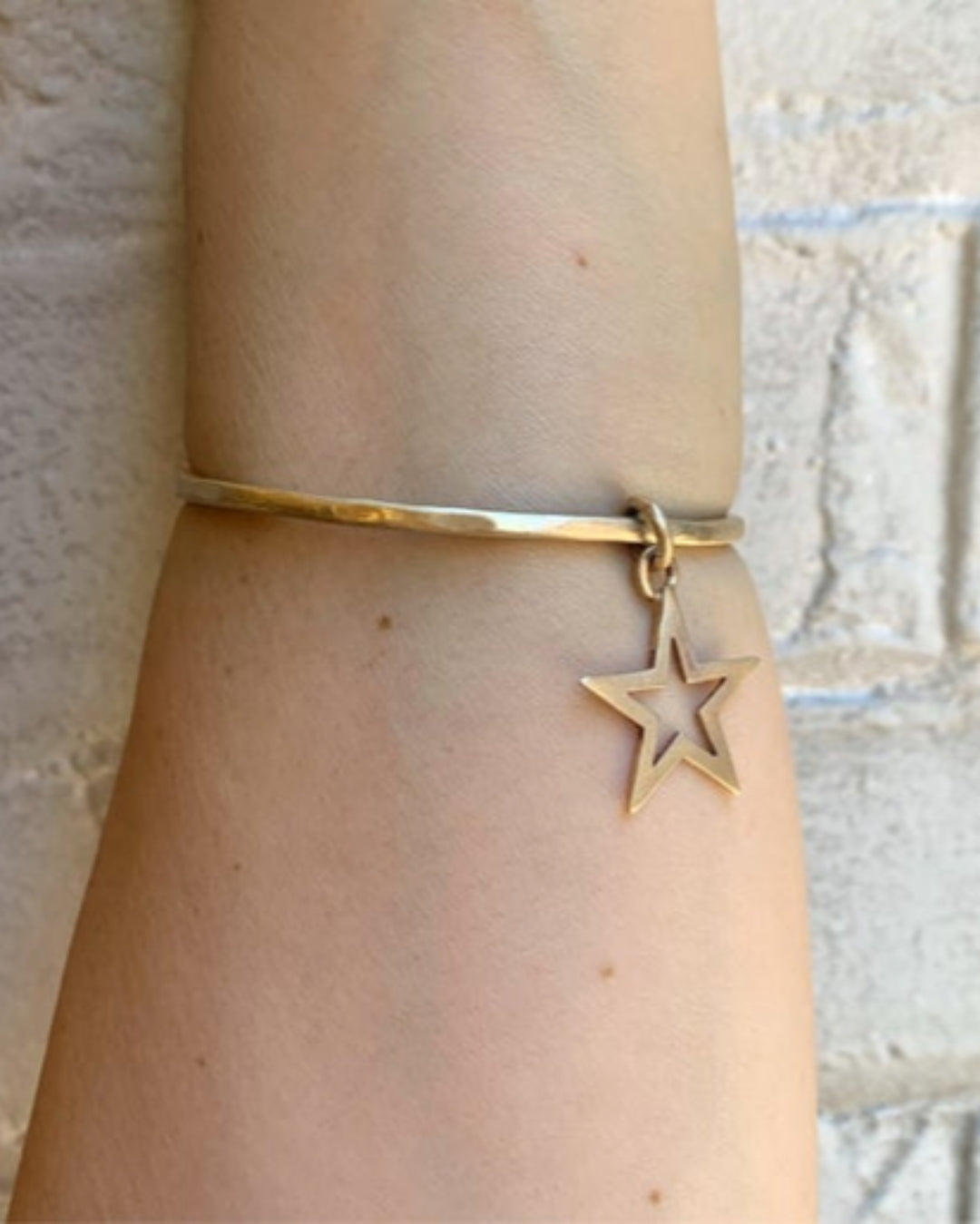 Gold bangle with a star charm on a wrist against a neutral background